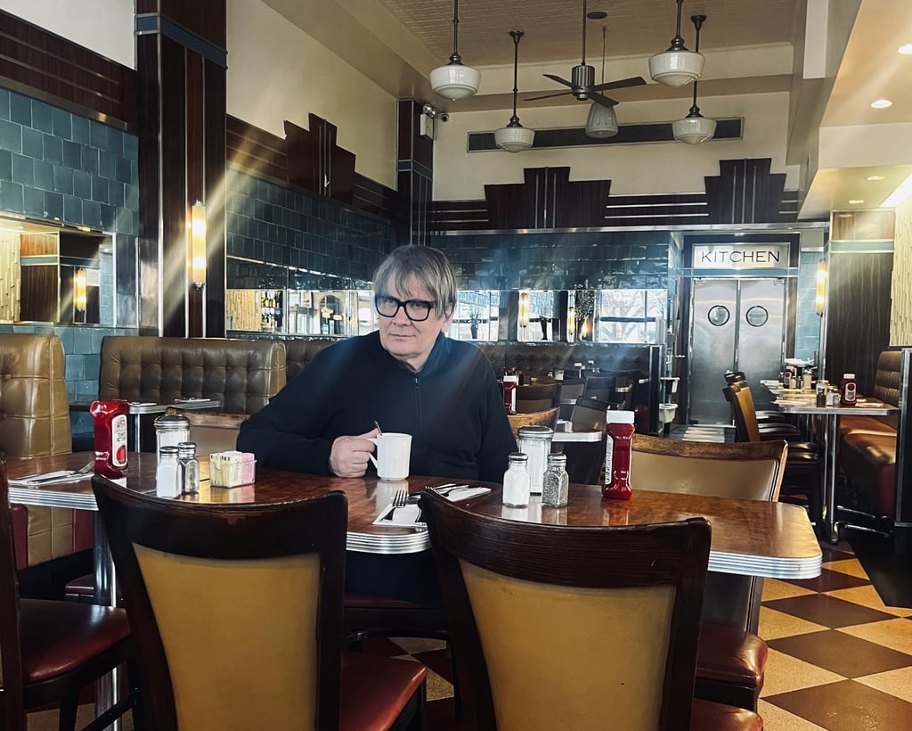 A person with short gray hair and glasses sits at a table in a retro diner, holding a white mug.