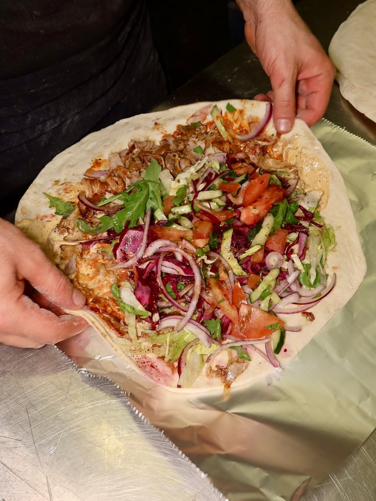 Hands assemble a flatbread wrap filled with grilled meat, lettuce, tomato, cucumber, red onion, and herbs.