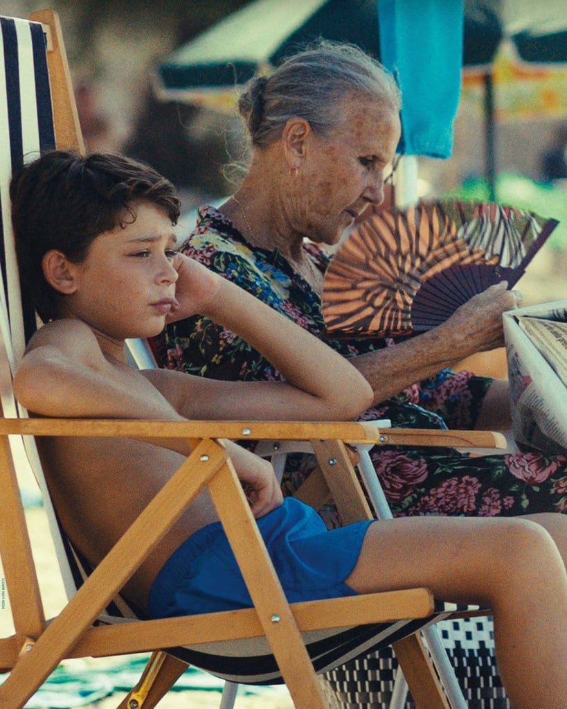 Woman and child sit in wooden beach chairs; the woman fans herself with a patterned folding fan while reading a magazine.