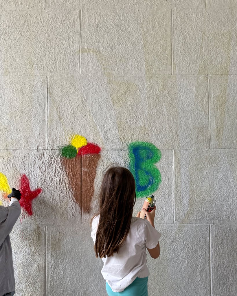 Two kids painting a mural on a gray brick wall with spray cans; bright circular shapes in yellow red green and blue visible on the wall behind them.] ,