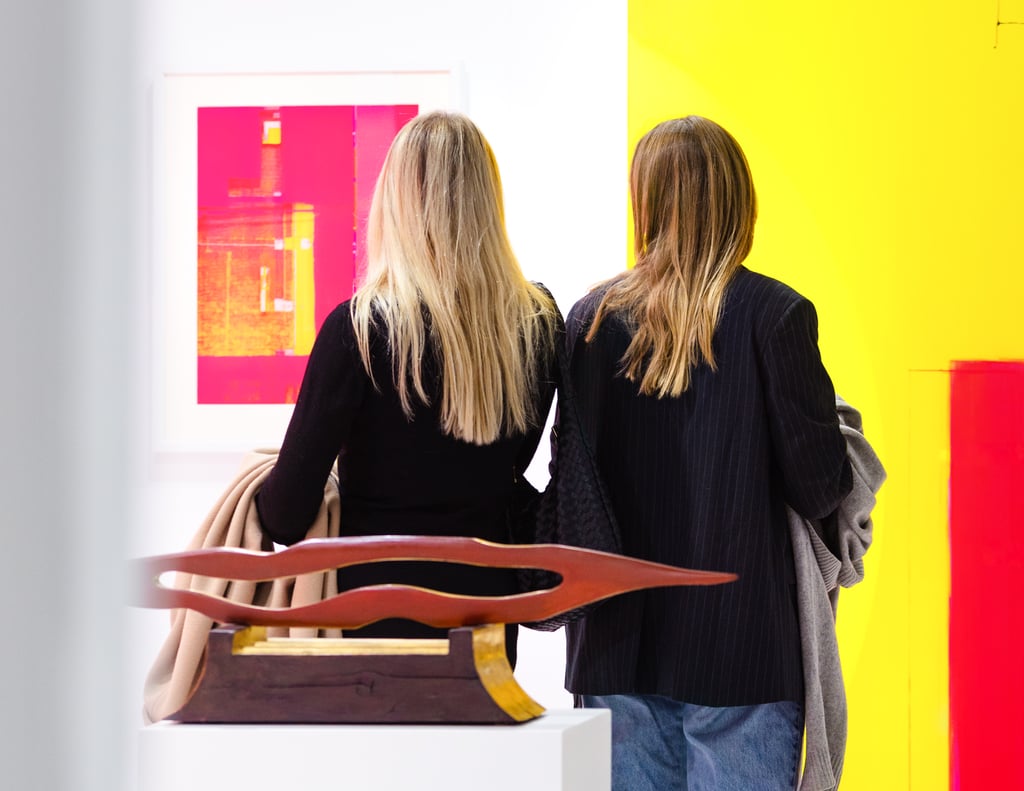 Two women seen from behind in a bright gallery, facing a pink abstract artwork on a white wall with a wooden sculpture in the foreground.