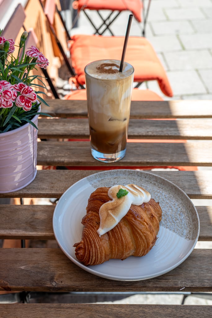 Iced coffee in a tall glass with straw on a wooden table, a croissant on a plate nearby, and pink flowers in a pot beside it outdoors.