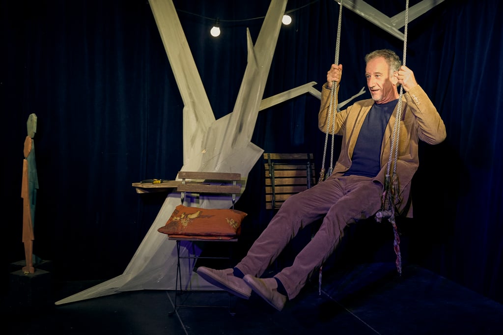 Theatrical performance: man in brown jacket on a rope swing prop against a dark stage backdrop.