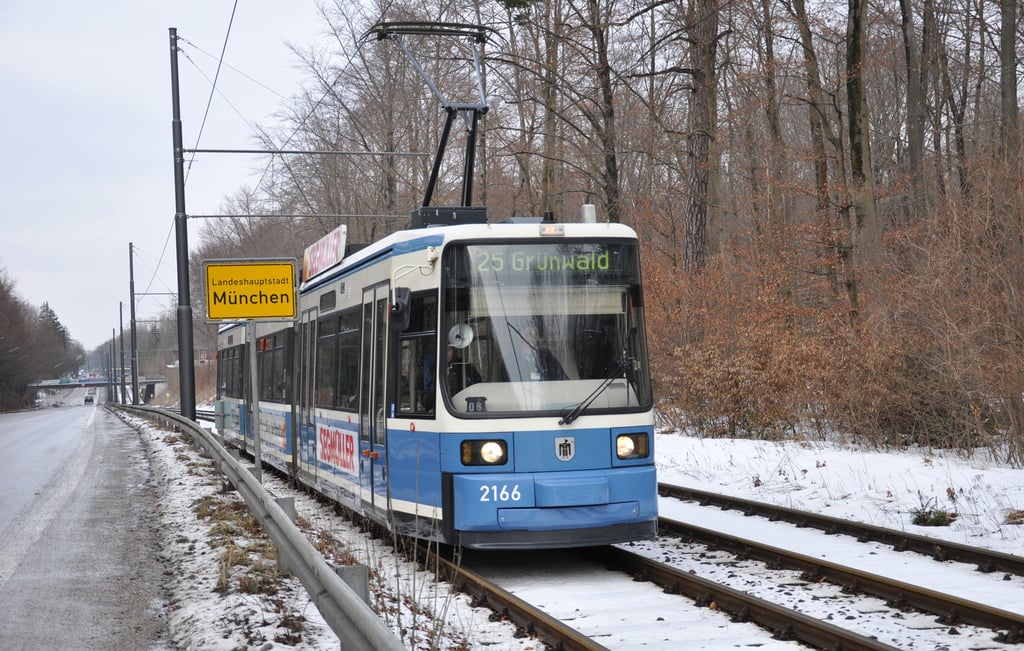 Tram number 2166 on a snow-covered track beside a road, with a yellow sign reading 'Landeshauptstadt München' and a forest in the background.