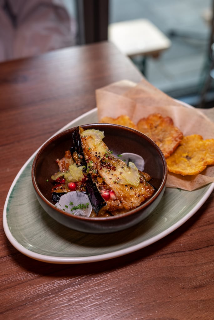 Bowl of roasted vegetables with pomegranate seeds and herbs, served with crispy chips on a parchment-lined side.
