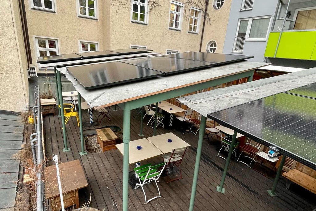 Outdoor courtyard with a covered seating area and solar panels on the roof; tables and chairs beneath green posts.