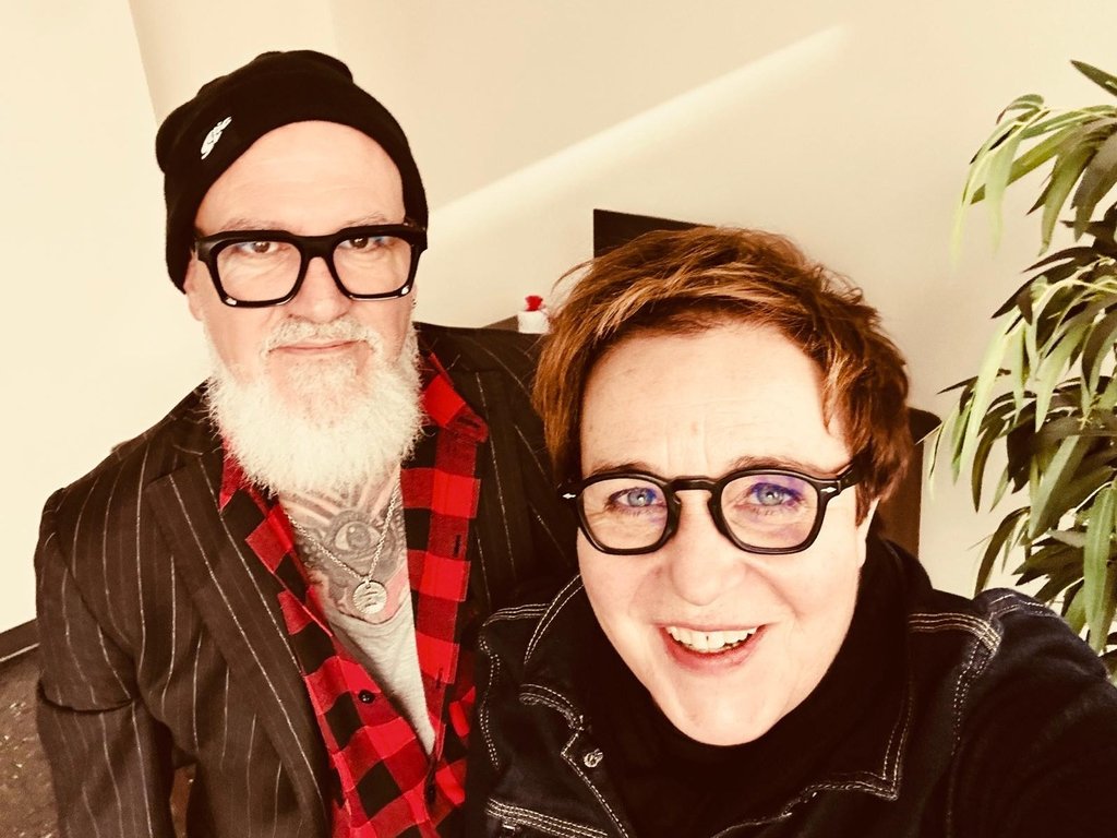 Two friends posing for a selfie indoors; man with black beanie, white beard, glasses, red plaid shirt and pinstripe jacket, beside a smiling woman with short hair and glasses.
