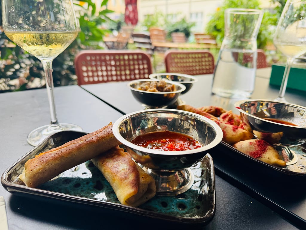 Outdoor dining scene with tapas: bowls of sauce, breadsticks, and spring rolls on a glass-tinished tray; blurred chairs and greenery in background.