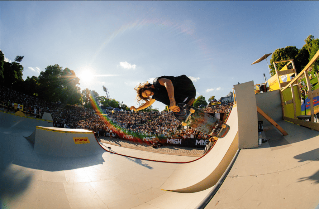 Skateboarder mid-air performing a trick over a halfpipe during a sunny outdoor competition, with a large cheering crowd and rainbow lens flare in the sky.