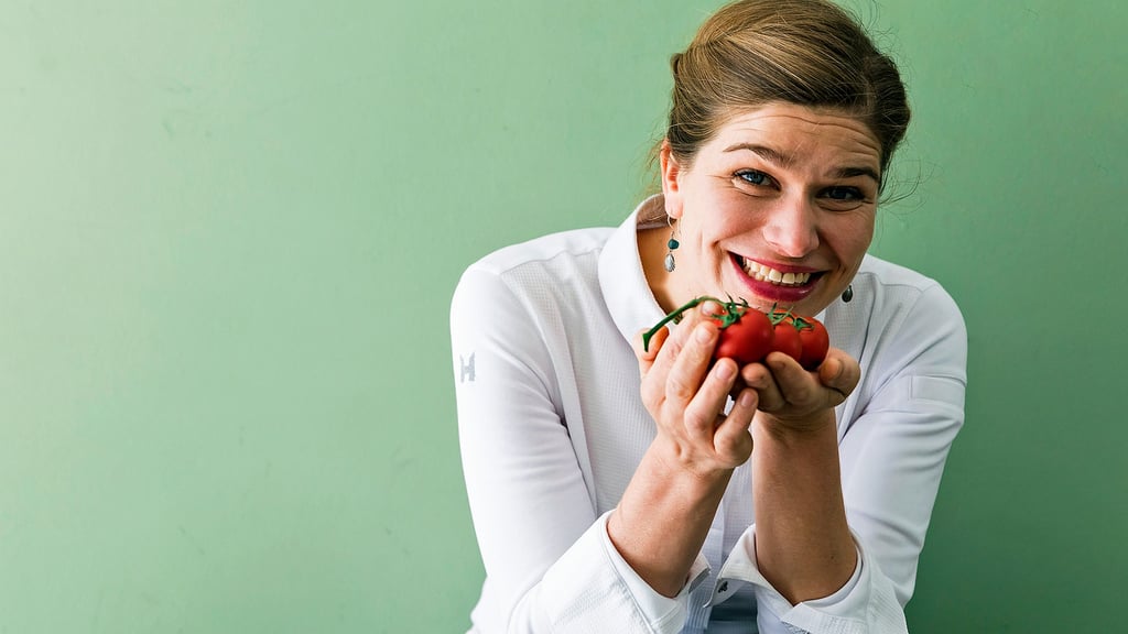 Smiling woman in a white shirt holds a bunch of ripe tomatoes toward the camera against a green wall