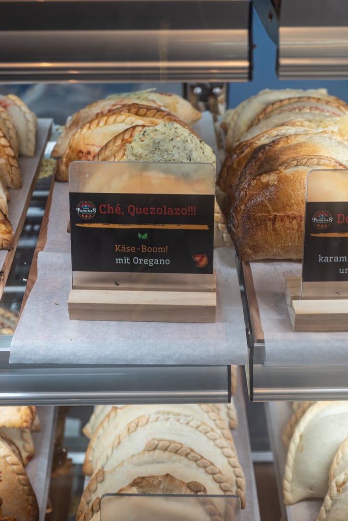Assorted crusty bread loaves in a bakery display with a label reading Käse‑Boom! mit Oregano.