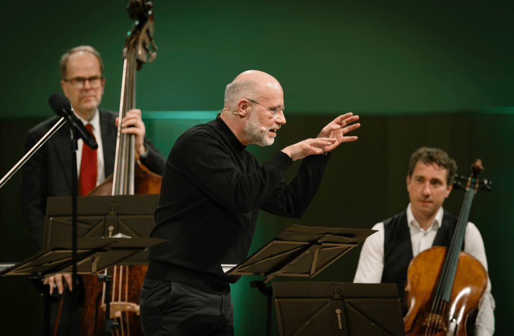 Conductor with headset leads a small string ensemble on stage, gesturing with hands while musicians play in the background.