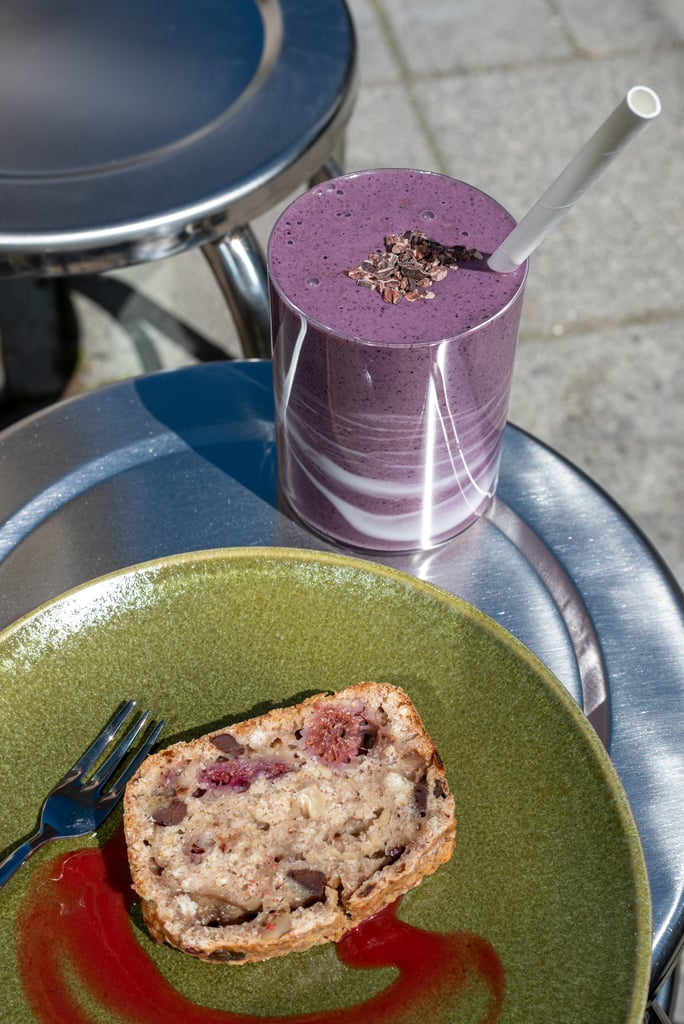 Purple berry smoothie in a tall glass with a white straw and cacao nibs, on a stack of plates with a slice of cake beside it outside
