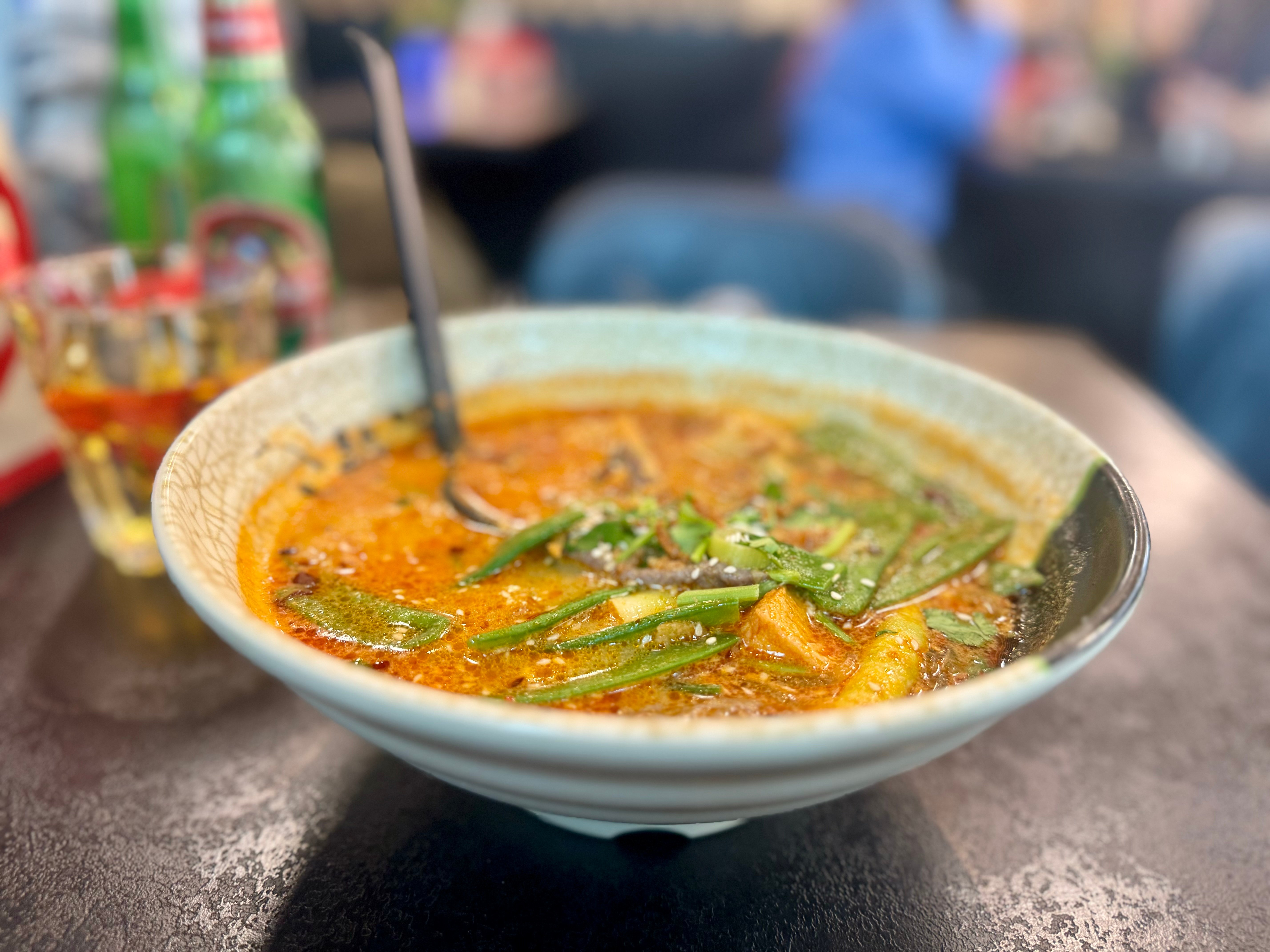 Schüssel Malatang-Suppe mit orangefarbener Brühe, Zuckerschoten und Kräutern auf einem dunklen Tisch