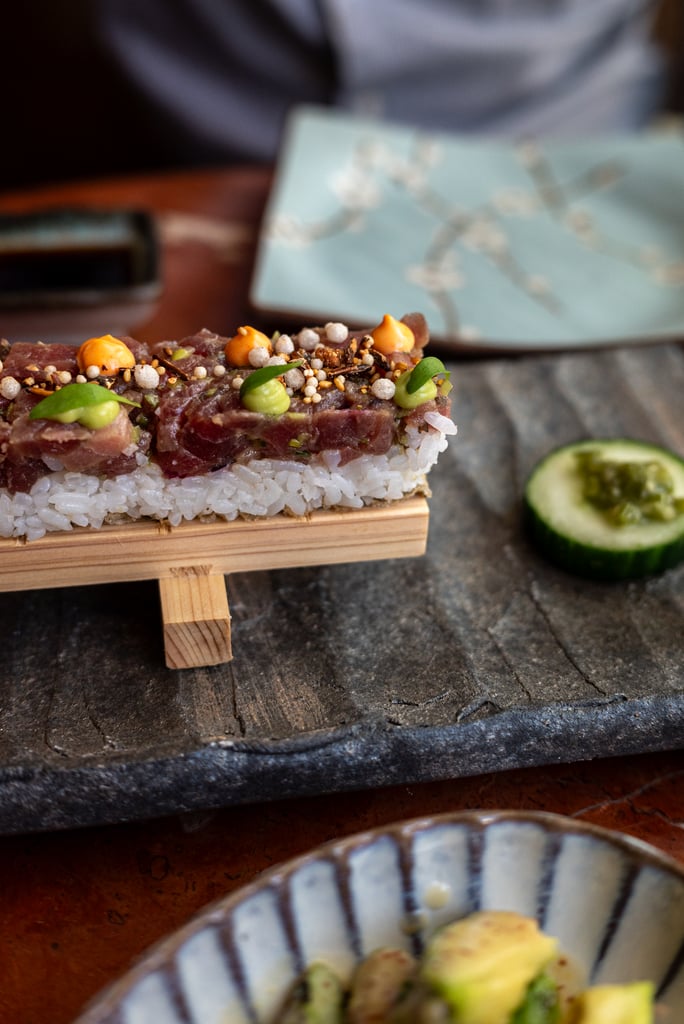 A layered sushi bite on a small wooden stand, with rice base, tuna slices, orange and green sauces, sesame pearls, and cucumber in the background.