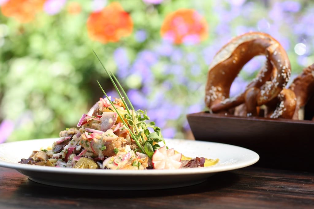 Plate of colorful chopped salad with herbs on a wooden table; blurred garden flowers and a pretzel basket in the background.