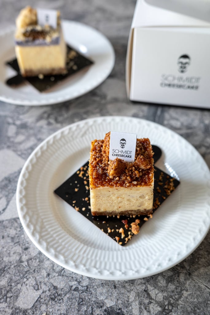 Close-up of a vanilla cheesecake square with caramel crumb topping on a white plate, small Schmidt Cheesecake label on top.