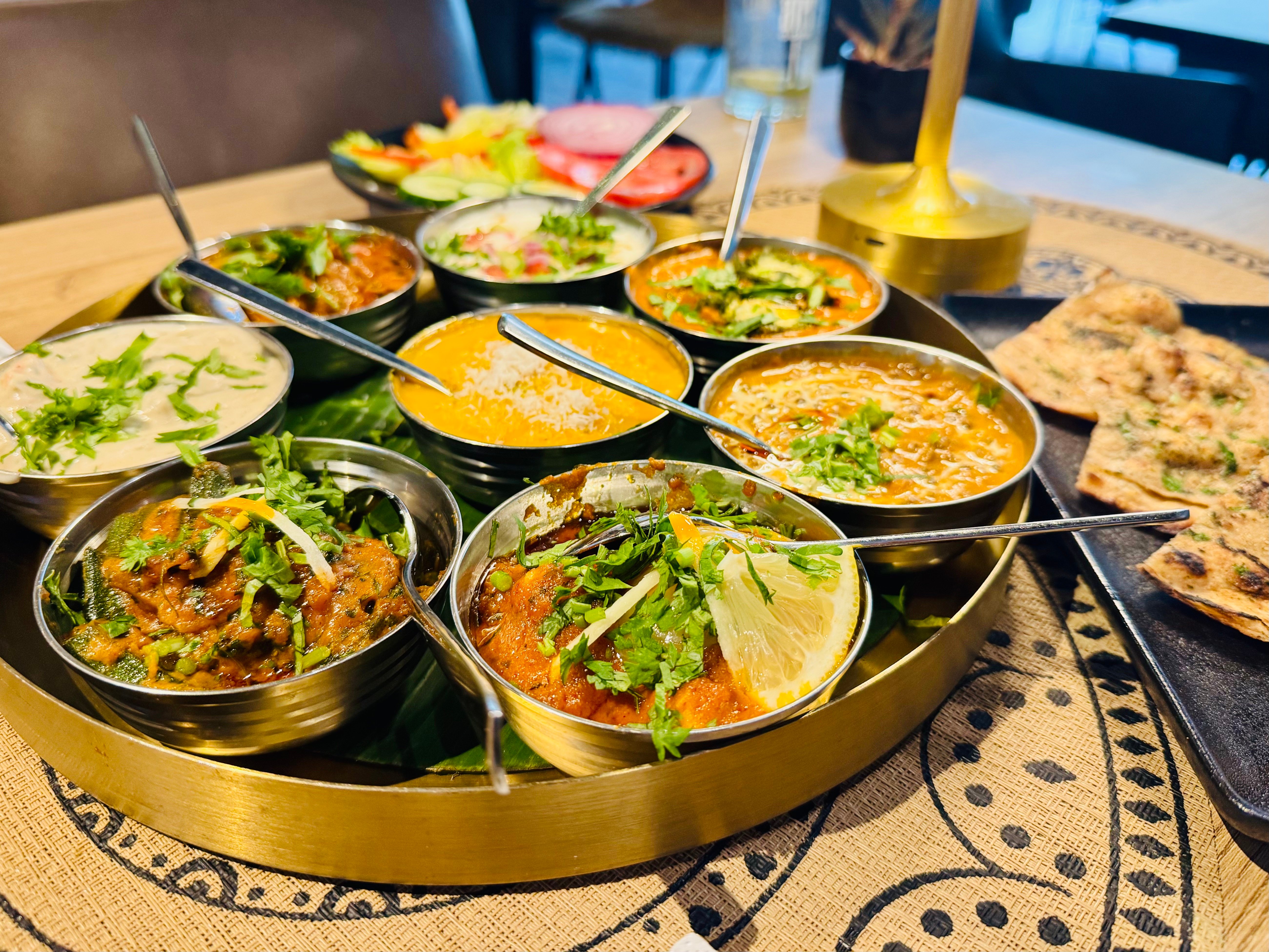 Thali-Tablett von oben mit Schälchen voller Curry, gelbem Daal und Raita, dazu Fladenbrot und Salat