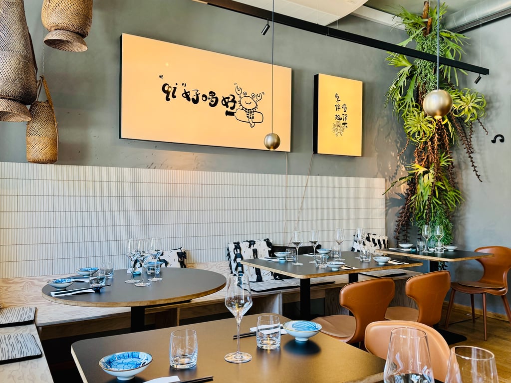 Modern restaurant dining area with round tables set with blue-patterned bowls, glasses, and tan chairs against a gray wall with wall art and greenery.