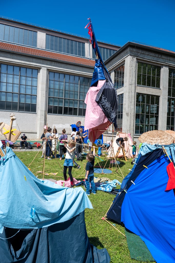 Festival scene with people assembling colorful fabric tents and poles outdoors near a modern building on a sunny day