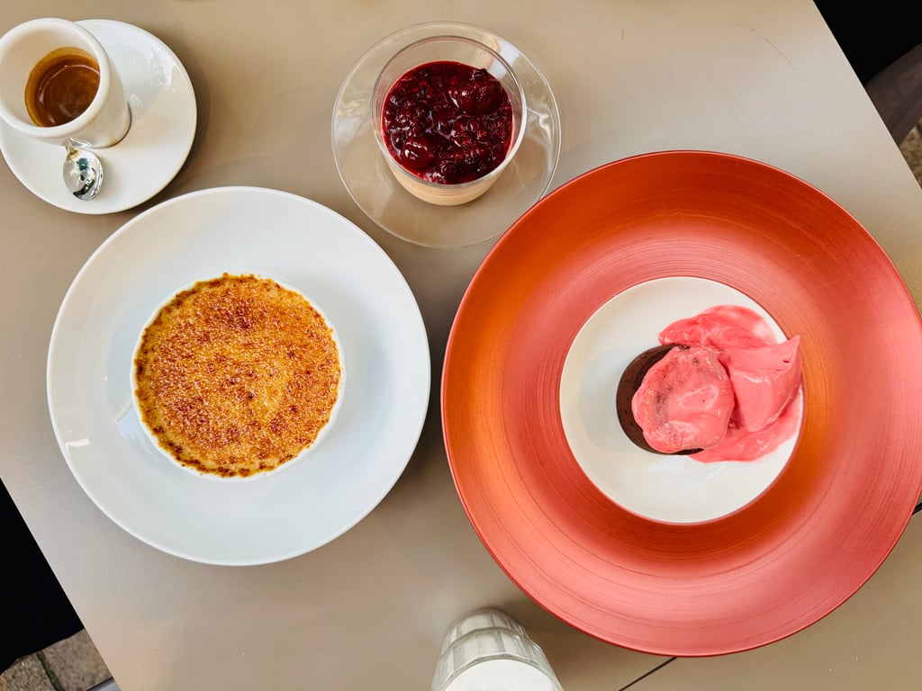 Top-down view of a dessert plate setup: crème brûlée on white plate, berry compote in a small glass, and pink ice cream scoops on an orange charger plate.