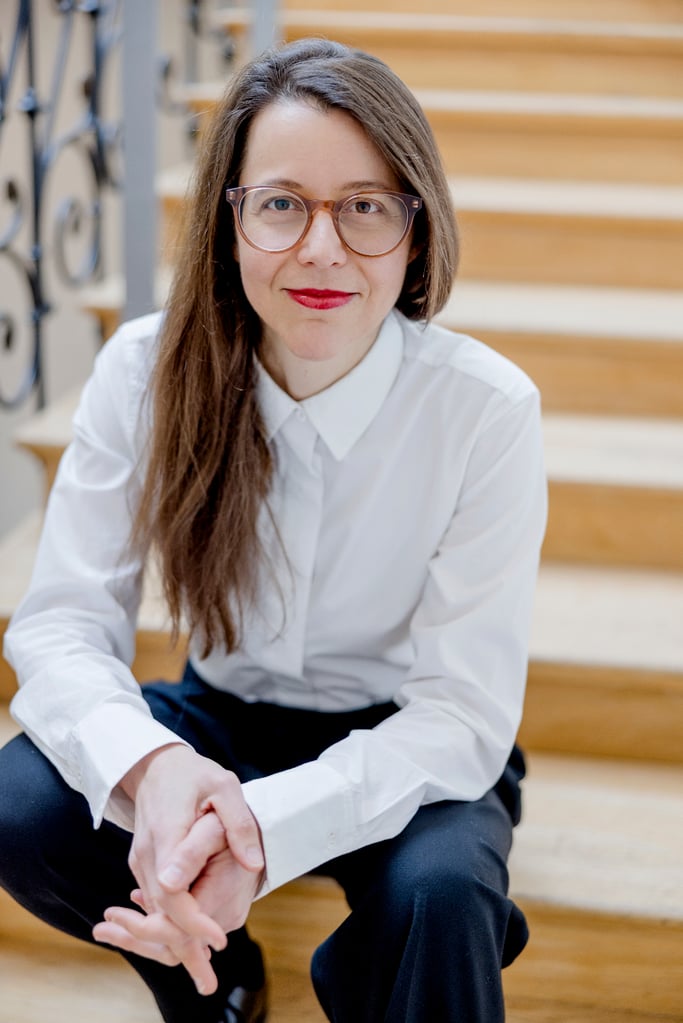 Woman with long brown hair and clear-framed glasses sits on wooden stairs, hands folded in her lap, wearing a white button-up shirt.