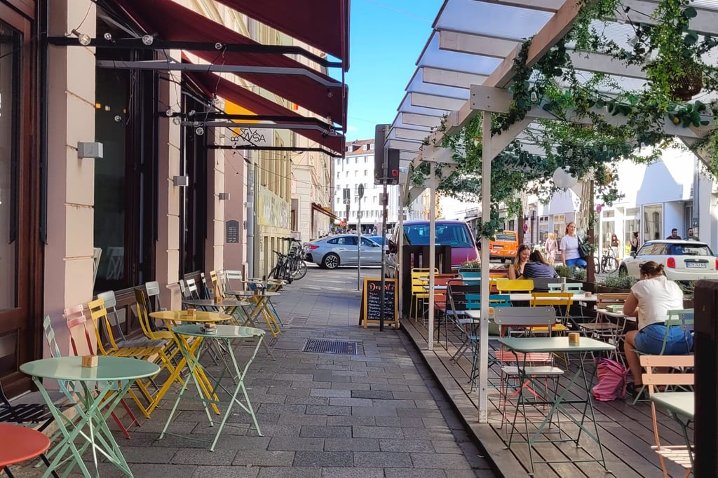 Outdoor cafe seating with colorful pastel chairs and tables along a pedestrian street under shade structures and string lights.] ,