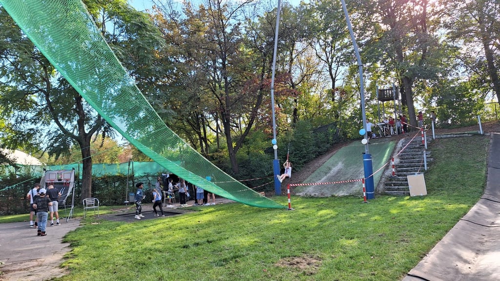 Children and adults gather at a park ropes course under a green safety net, with trees and stairs nearby on a sunny day.
