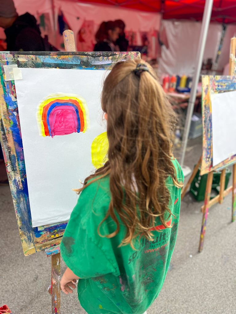 Child in a green shirt painting a colorful rainbow on a white canvas at an outdoor art booth.