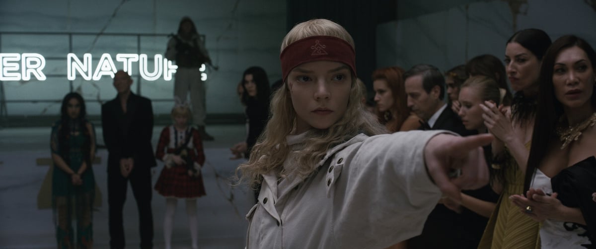 Anya Taylor-Joy mit rotem Stirnband zeigt in einer Szene aus „Sacrifice“ mit ausgestrecktem Arm nach vorn, hinter ihr eine Gruppe von Gala-Gästen.