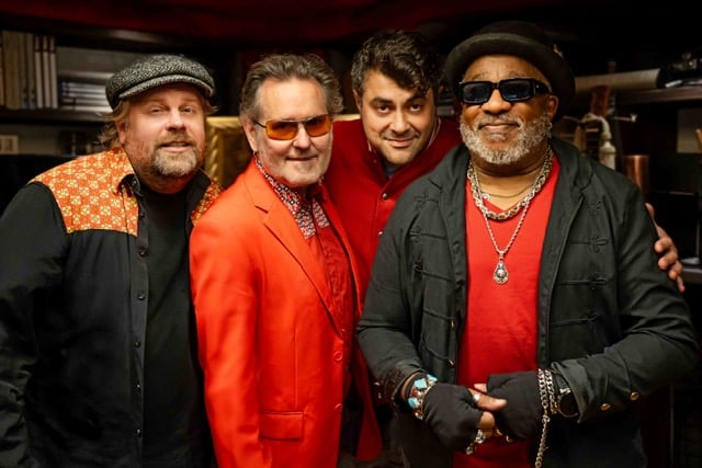 Four men posing together in a backstage/club setting, wearing bold jackets and sunglasses for a group photo.