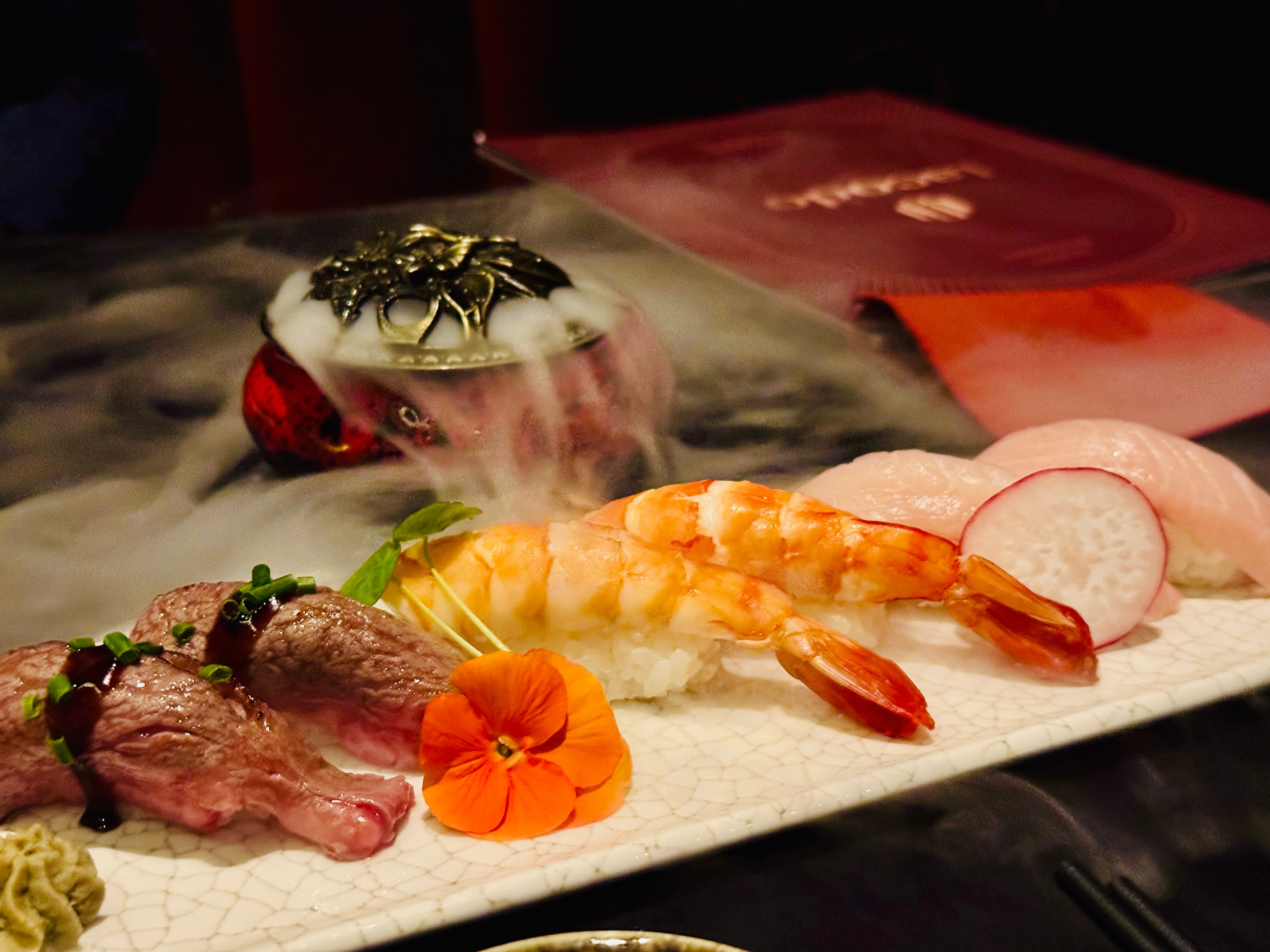 Nigiri mit Wagyu, Garnelen und Rettichscheibe auf heller Platte, dahinter eine Schale mit aufsteigendem Trockeneis-Nebel