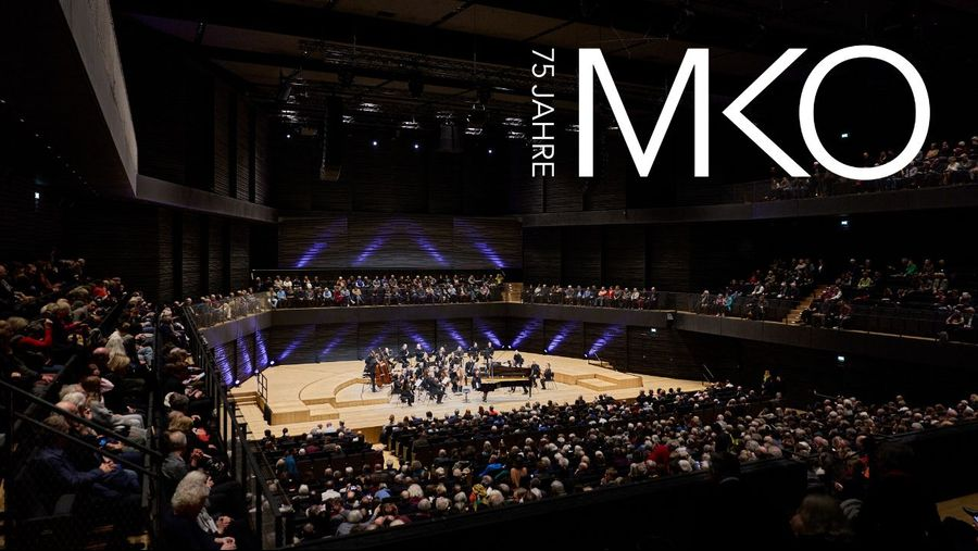Large modern concert hall filled with audience watching an orchestra perform on a circular stage, MKO logo in the upper-right corner with '75 Jahre' text alongside.
