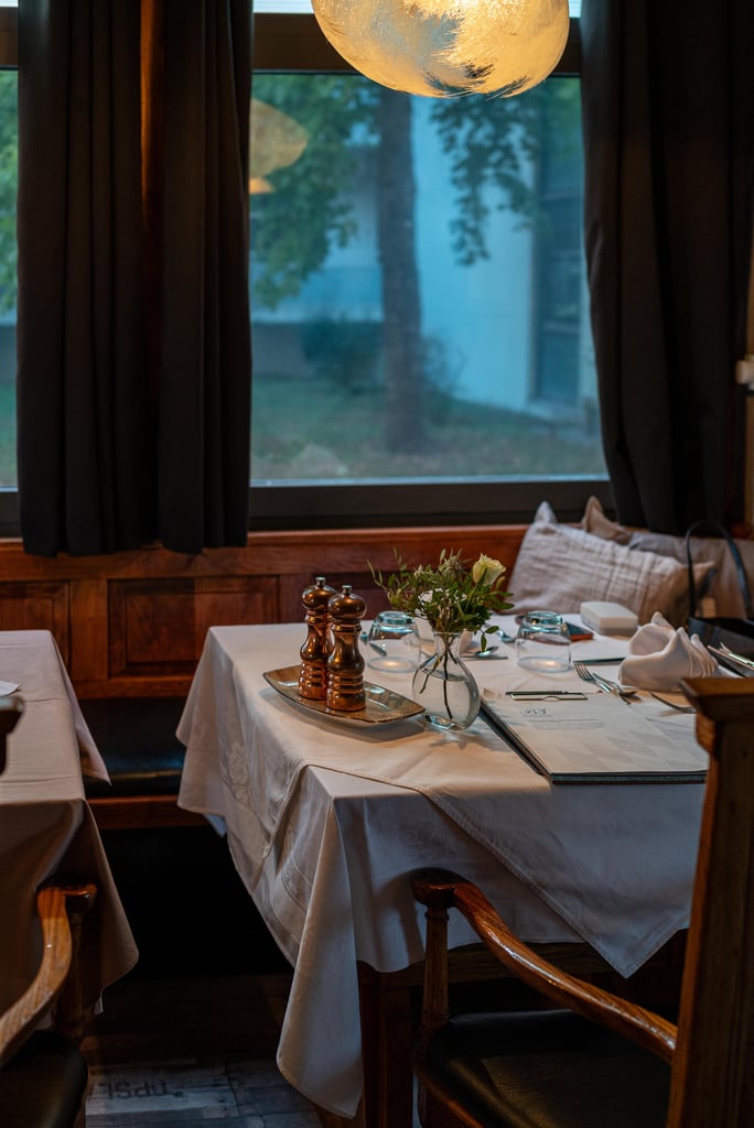 Formal dining table set with white tablecloth, glassware, folded napkins, and a small flower vase in a cozy restaurant.","Table for dining: white linens, silverware, and a centerpiece vase, against a window with dark curtains.","Elegant restaurant table with place settings, pepper grinders, and a tiny bouquet, near a window.","Intimate dining setup featuring a white-clothed table, candles, and a small floral arrangement.","Close-up of a dining table with utensils, glasses, and a vase, in a warmly lit dining room.