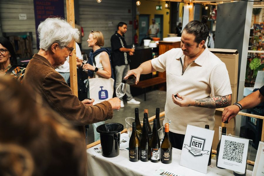Wine tasting at an outdoor market: a staff member pours wine into a glass for an older man, bottles lined up on the table in front of them.