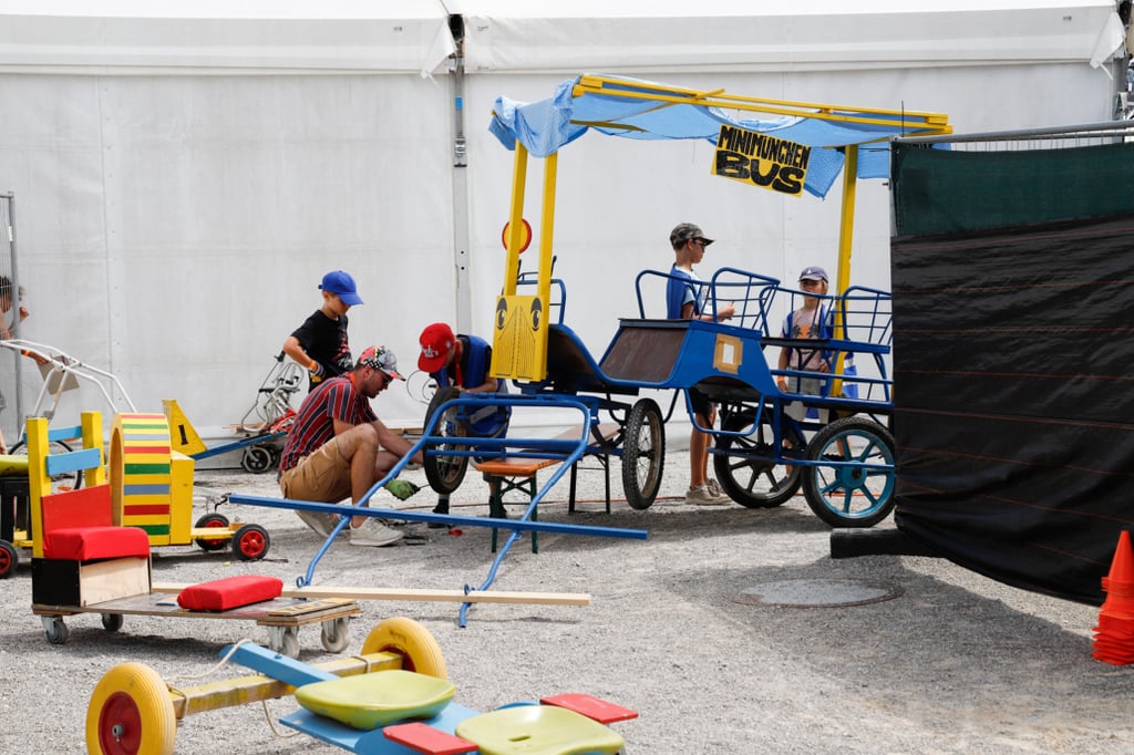A man crouches beside a blue wheeled ride with children standing on it at a fairground setup and a white tent in the background.