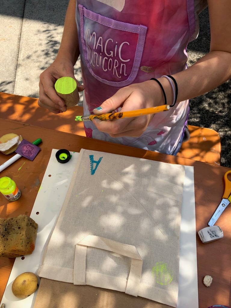 Child in a pink apron painting a cork neon green on an outdoor craft table, holding a paintbrush nearby.