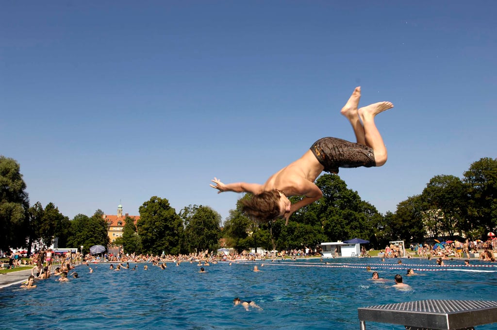 Person flipping into a crowded outdoor pool on a sunny day, mid-dive with legs in the air.