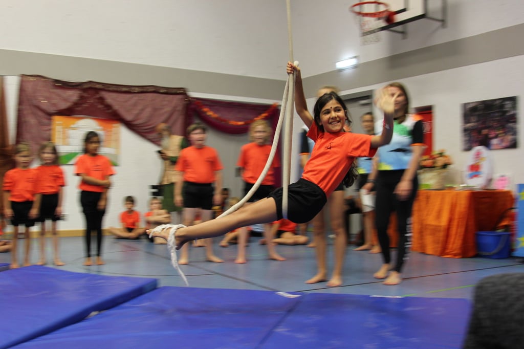 Girl in an orange shirt swinging on a rope in a gym, with classmates watching in the background.