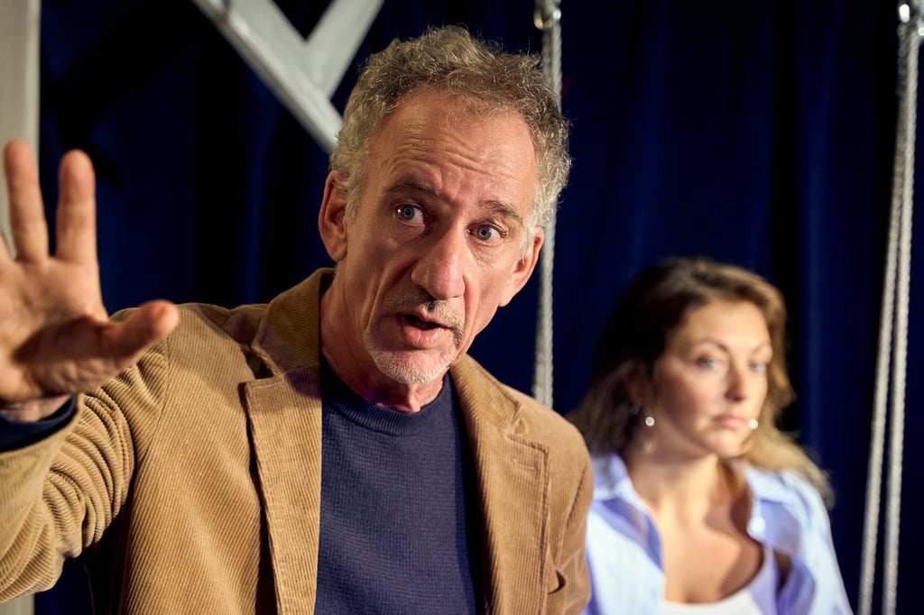 Man in a brown blazer raises his left hand while speaking; two people sit in the background against a dark blue curtain.