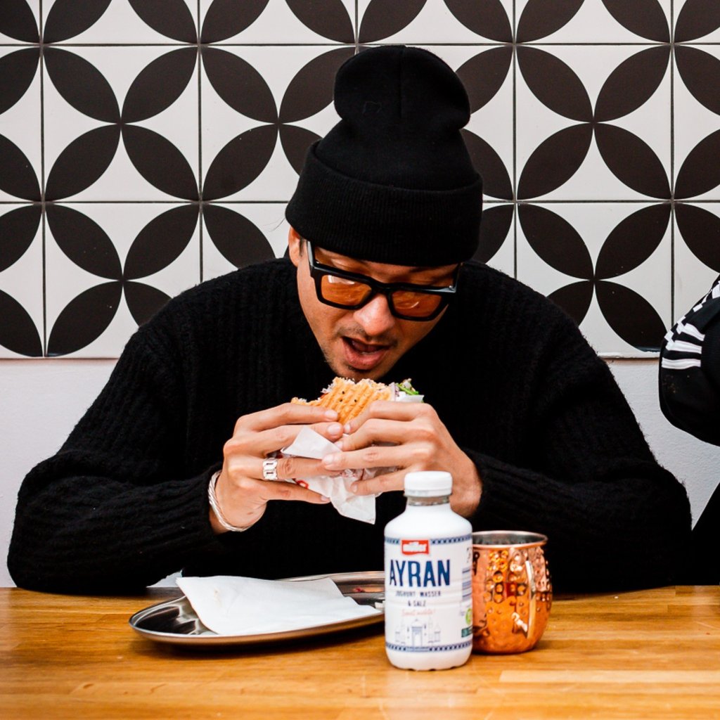 Man in a black beanie and glasses bites into a sandwich at a wooden table, patterned wall behind him.