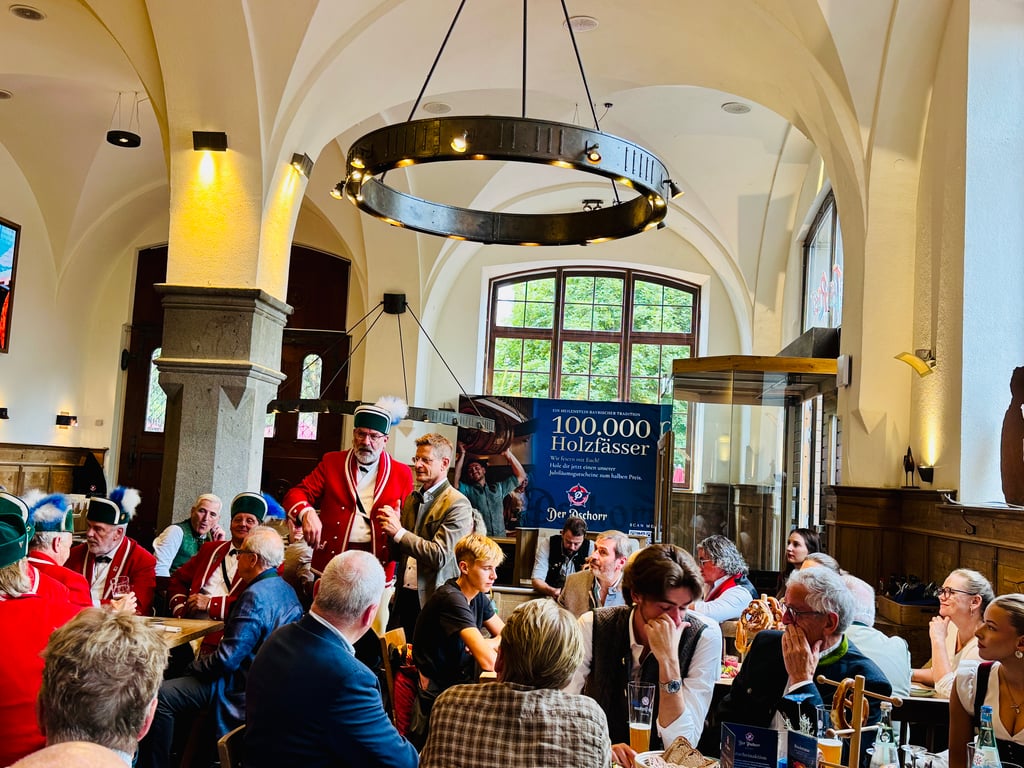 Group of people in traditional Bavarian clothing sharing a meal and drinks in a bright hall with arched ceilings and warm lighting, festive atmosphere.