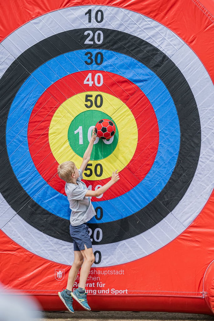 Boy reaches for a red-and-black soccer ball in front of a giant inflatable archery target with colorful rings and numbers 10–50.