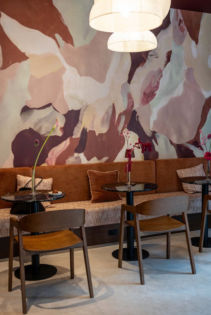 Cafe seating area with brown upholstered bench, wooden chairs, round black tables, and pink flowers against a pastel abstract mural.