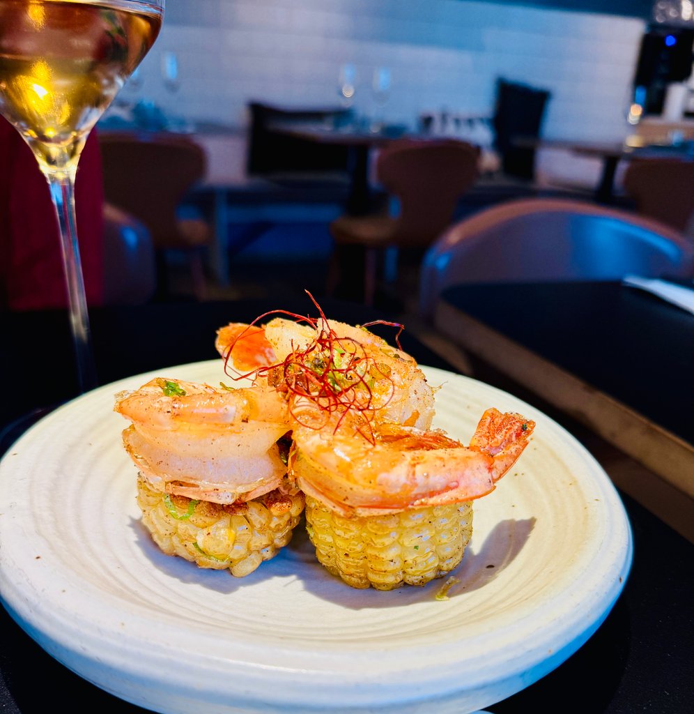 Stacked shrimp on rice cakes with herbs on a white plate, a wine glass in the foreground in a dim restaurant setting.