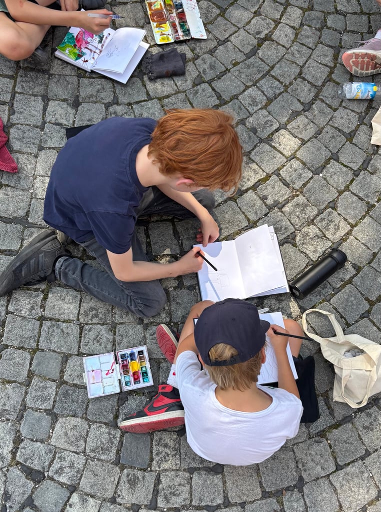 Group of children drawing and painting on a cobblestone pavement with watercolor palettes and sketchbooks nearby.