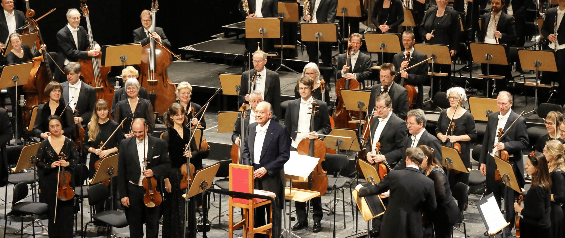 Full orchestra in formal black attire on stage after a performance, with string players holding instruments and seated stands in the background.
