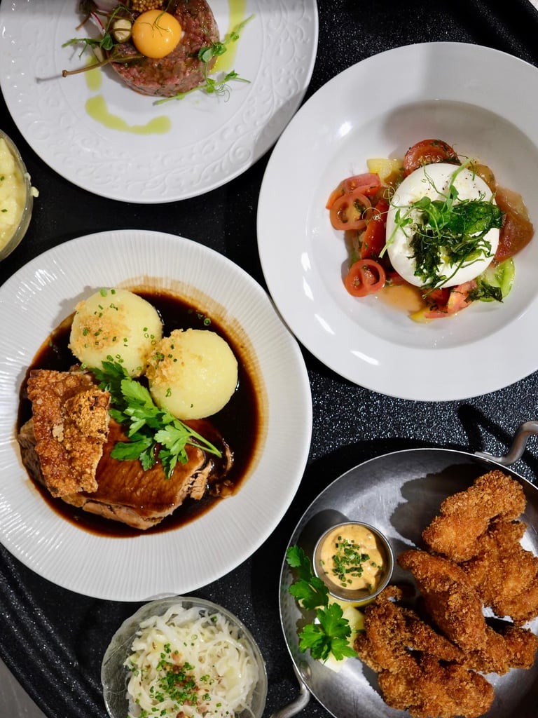 Assorted dishes on a black table: beef with croquettes and gravy, burrata with tomato and greens, fried chicken bites with dipping sauce, and creamy pasta sides.