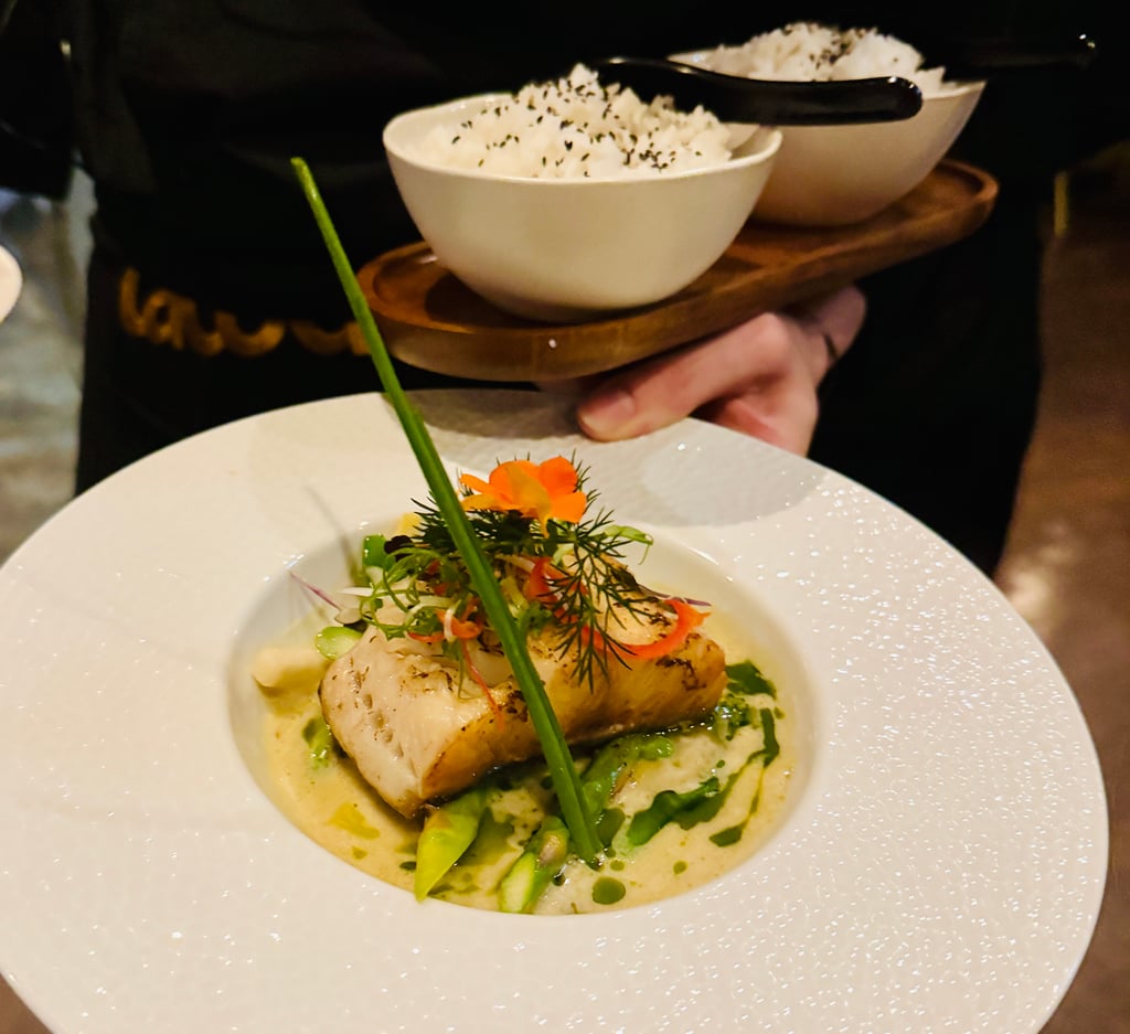 Plate with seared fish fillet over green sauce and herbs, orange flower garnish, and white rice with sesame in the background on a wooden board.