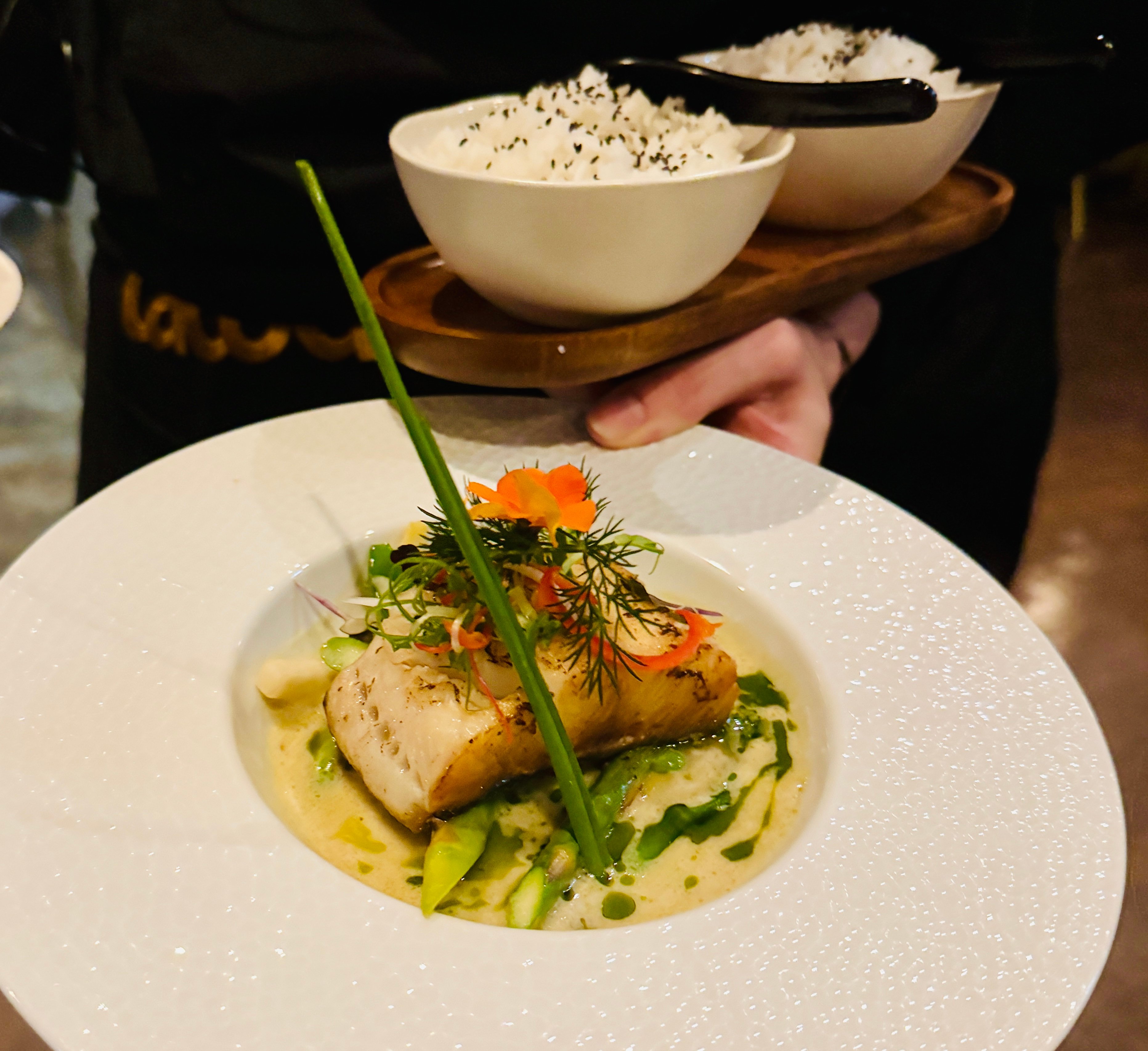 Plate with seared fish fillet over green sauce and herbs, orange flower garnish, and white rice with sesame in the background on a wooden board.